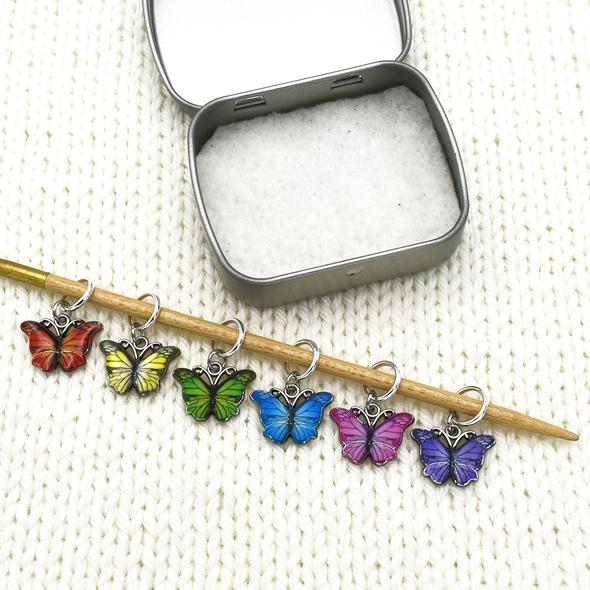 Set of six enamel butterfly charm snag free ring stitch markers in red, yellow, green, blue, pink and purple on knitting needle resting on white knitted square with storage tin for knitting by Pretty Warm Designs