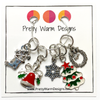 Five silver alloy and enamel Christmas themed snag free ring stitch markers for knitting attached to silver hoop on cardstock with Pretty Warm Designs logo
