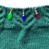 Green, blue and red faux Christmas lights knitting stitch markers threaded on a red knitting needle cable laying on a green knitting background sold by Pretty Warm Designs Inc.