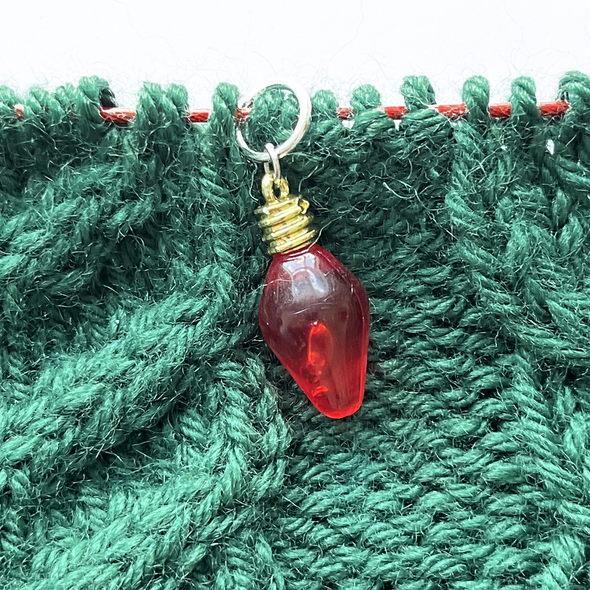 One red faux Christmas light knitting stitch marker threaded on a red knitting needle cable laying on a green knitting background sold by Pretty Warm Designs Inc.