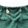 Two winter themed enamel stitch markers for knitting threaded on knitting needle cable and laying on green knitting sold by Pretty Warm Designs Inc