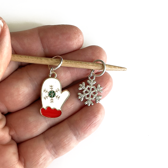 Two Winter-themed knitting ring stitch markers, one red, white and green enamel mitten and one silver toned snowflake with rhinestones on a bamboo knitting needle held on a hand, made and sold by Pretty Warm Designs