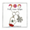 Two Winter-themed knitting ring stitch markers, one red, white and green enamel mitten and one silver toned snowflake with rhinestones attached with a silver hoop ring to cardstock with Pretty Warm Designs text and logo
