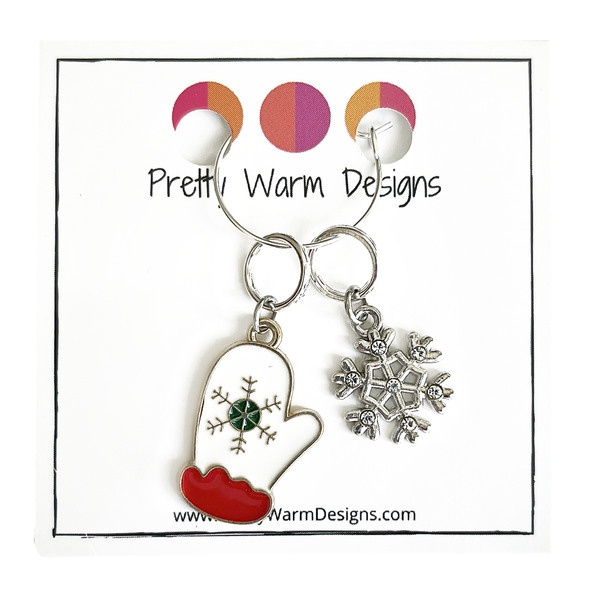 Two Winter-themed knitting ring stitch markers, one red, white and green enamel mitten and one silver toned snowflake with rhinestones attached with a silver hoop ring to cardstock with Pretty Warm Designs text and logo