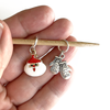 One red and white enamel Santa stitch marker and one silver alloy mittens stitch marker on a bamboo needle for knitting held on a hand, made and sold by Pretty Warm Designs