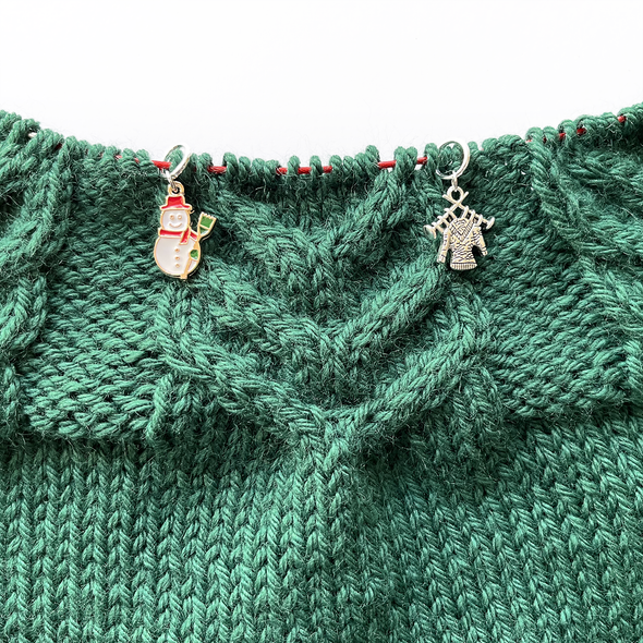 Two winter themed enamel stitch markers for knitting threaded on knitting needle cable and laying on green knitting sold by Pretty Warm Designs Inc