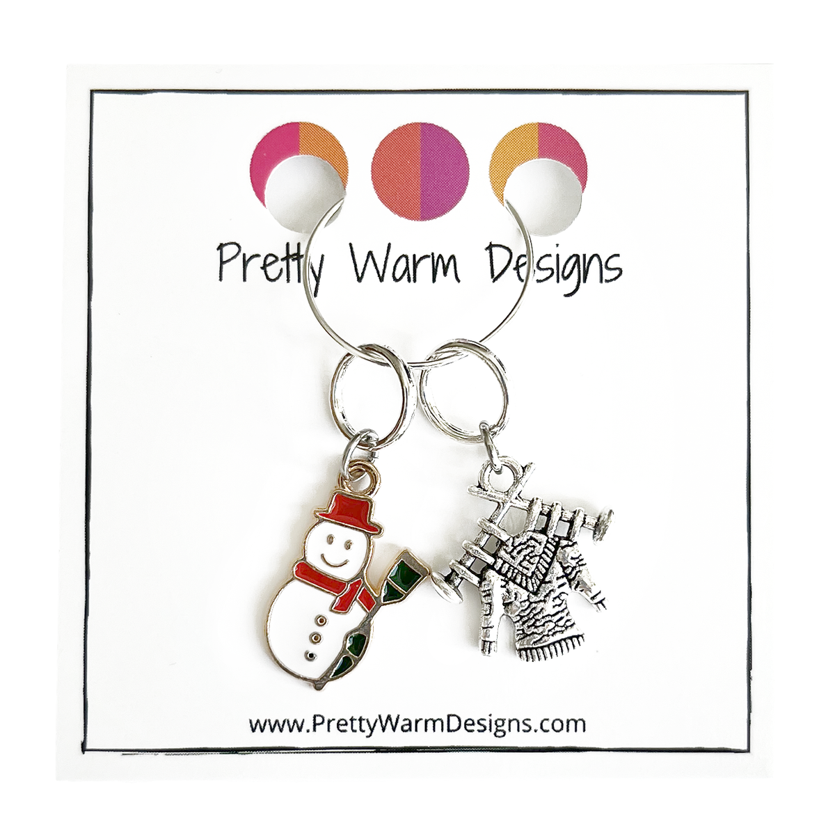 Two Winter-themed knitting ring stitch markers, one red, white and green enamel snowman and one antiqued Tibetan silver sweater on knitting needles attached with silver hoop ring to cardstock with Pretty Warm Designs text and logo