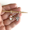 Two Winter-themed knitting ring stitch markers, one red, white and green enamel snowman and one antiqued Tibetan silver sweater on knitting needles held on bamboo needle on a hand, made and sold by Pretty Warm Designs
