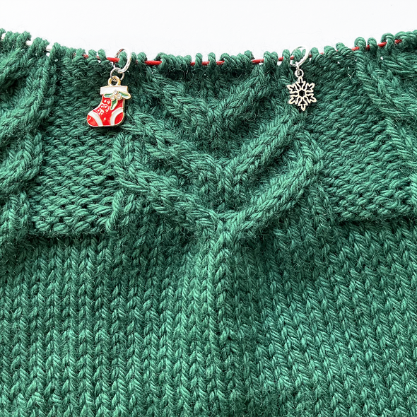 Two Christmas themed stitch markers for knitting threaded on knitting needle cable on top of green knitting sold by Pretty Warm Designs Inc.