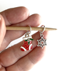One red, white and green enamel with rhinestones Christmas stocking ring stitch marker and one silver alloy snowflake ring stitch marker for knitting on a bamboo knitting needle held in a hand, made and sold by Pretty Warm Designs
