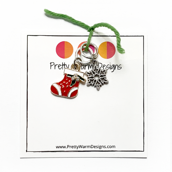 One red, white and green enamel with rhinestones Christmas stocking ring stitch marker and one silver alloy snowflake ring stitch marker for knitting tied with green yarn to cardstock with Pretty Warm Designs text and logo