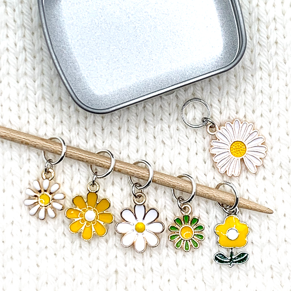 Set of six white, yellow and green enamel daisy charms snag free ring stitch markers on bamboo knitting needle resting on white knitted background with tin for knitting by Pretty Warm Designs