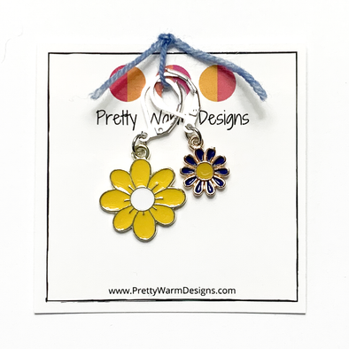 Two enamel charm crochet locking stitch markers with one yellow and one blue daisy attached with blue yarn to cardstock with Pretty Warm Designs text and logo