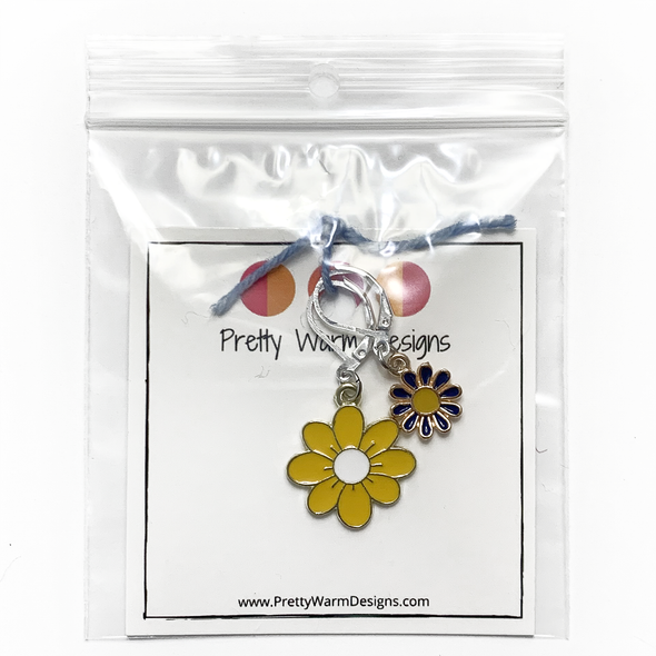 Two enamel charm crochet locking stitch markers with one yellow and one blue daisy attached with blue yarn to cardstock with Pretty Warm Designs text and logo packaged in clear poly bag