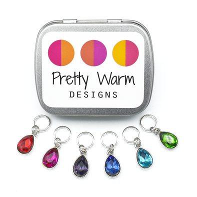Set of six coloured rhinestones in red, pink, purple, blue, turquoise and green set in silver toned snag free ring stitch markers for knitting and decorative tin with Pretty Warm Designs text and logo