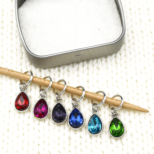 Set of six coloured rhinestones in red, pink, purple, blue, turquoise and green set in silver toned snag free ring stitch markers on a knitting needle and open decorative tin by Pretty Warm Designs