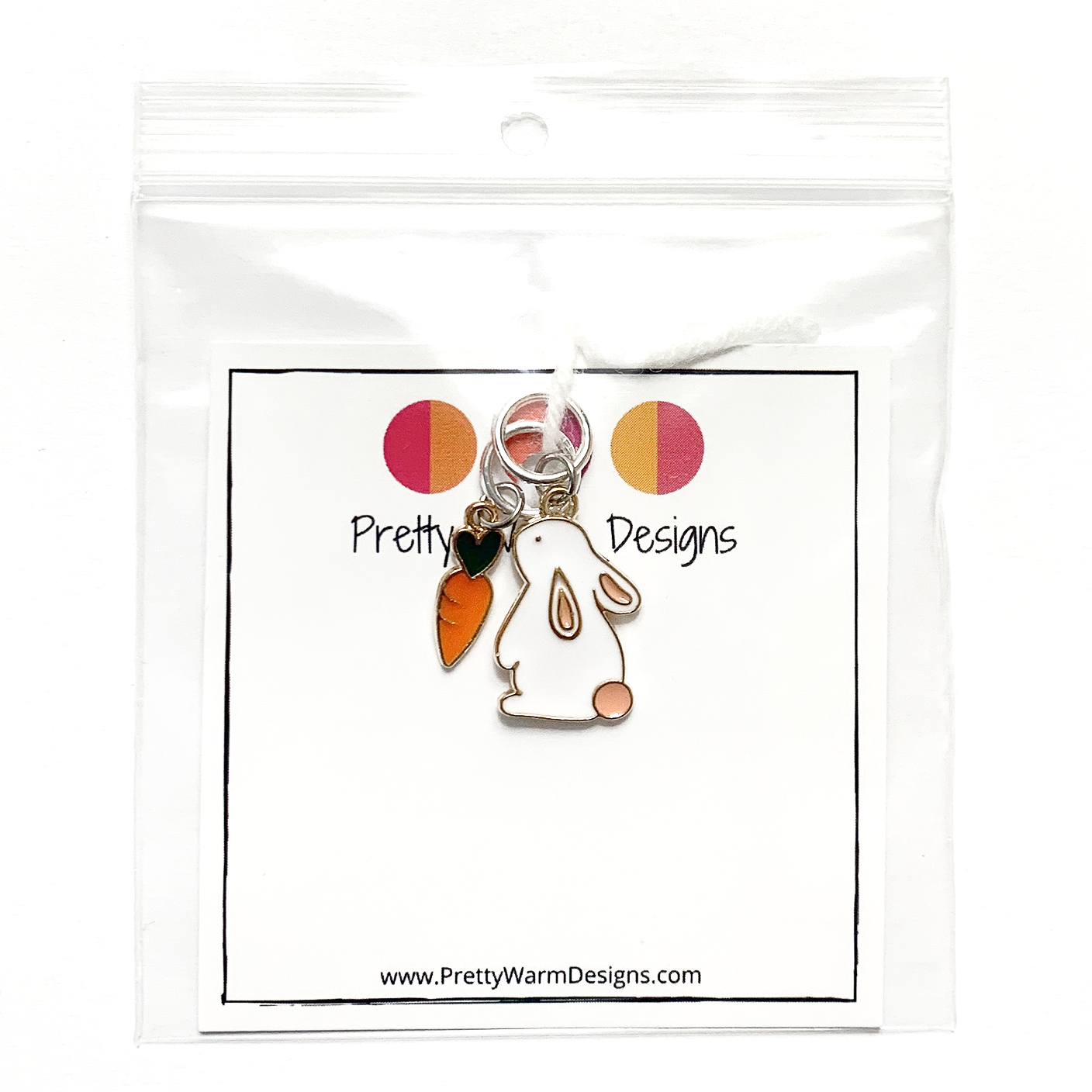 Two knitting ring stitch markers, one white and peach enamel bunny and one orange and green enamel carrot attached with white yarn to cardstock with Pretty Warm Designs text and logo packaged in a clear poly bag