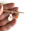Two knitting ring stitch markers, one white and peach enamel bunny and one orange and green enamel carrot on bamboo knitting needle held on a hand, made and sold by Pretty Warm Designs