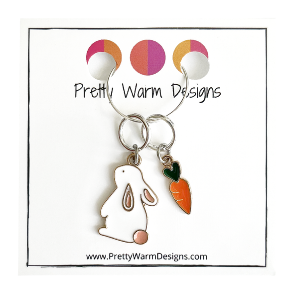 Two knitting ring stitch markers, one white and peach enamel bunny and one orange and green enamel carrot attached with silver hoop ring to cardstock with Pretty Warm Designs text and logo