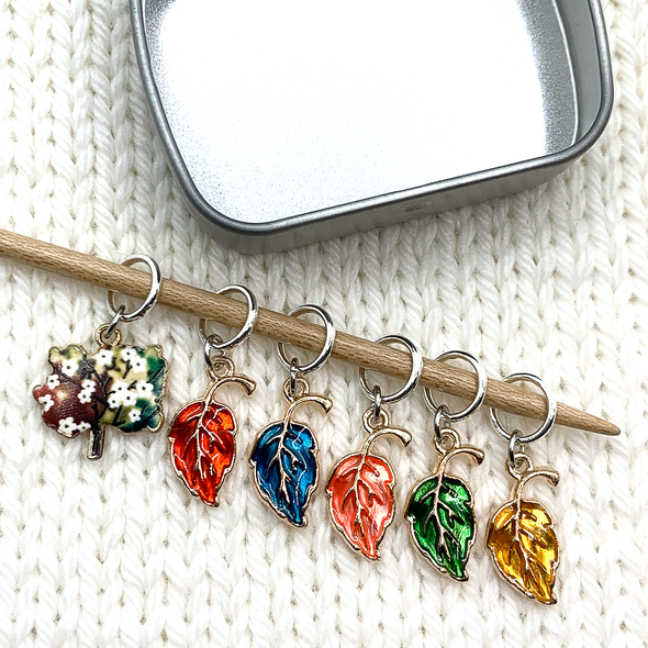 Set of five red, blue, orange, green and yellow enamel leaves and one tree charm snag free ring stitch markers on bamboo knitting needle resting on a white knitted background with decorative tin for knitting by Pretty Warm Designs