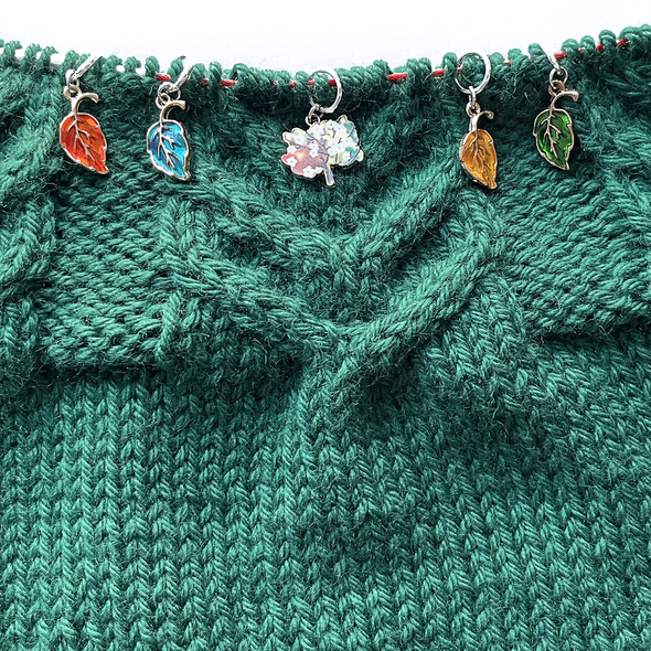 Four red, blue, orange and green enamel leaves and one tree charm snag free ring stitch markers threaded on a red knitting needle cable laying on a green knitted background and sold by Pretty Warm Designs Inc.