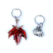 Two Fall-themed knitting ring stitch markers, one red enamel maple leaf and one silver toned pumpkin by Pretty Warm Designs