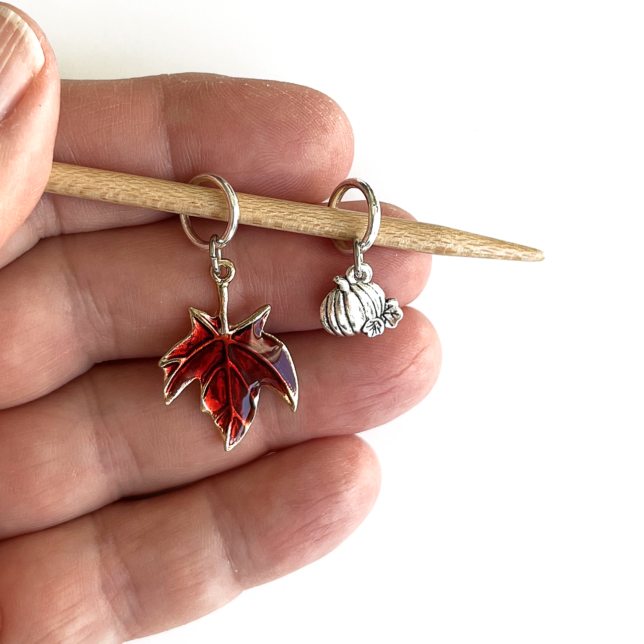 One red enamel maple leaf knitting stitch marker and one silver alloy pumpkin knitting stitch marker on a bamboo knitting needle held in a hand, made and sold by Pretty Warm Designs