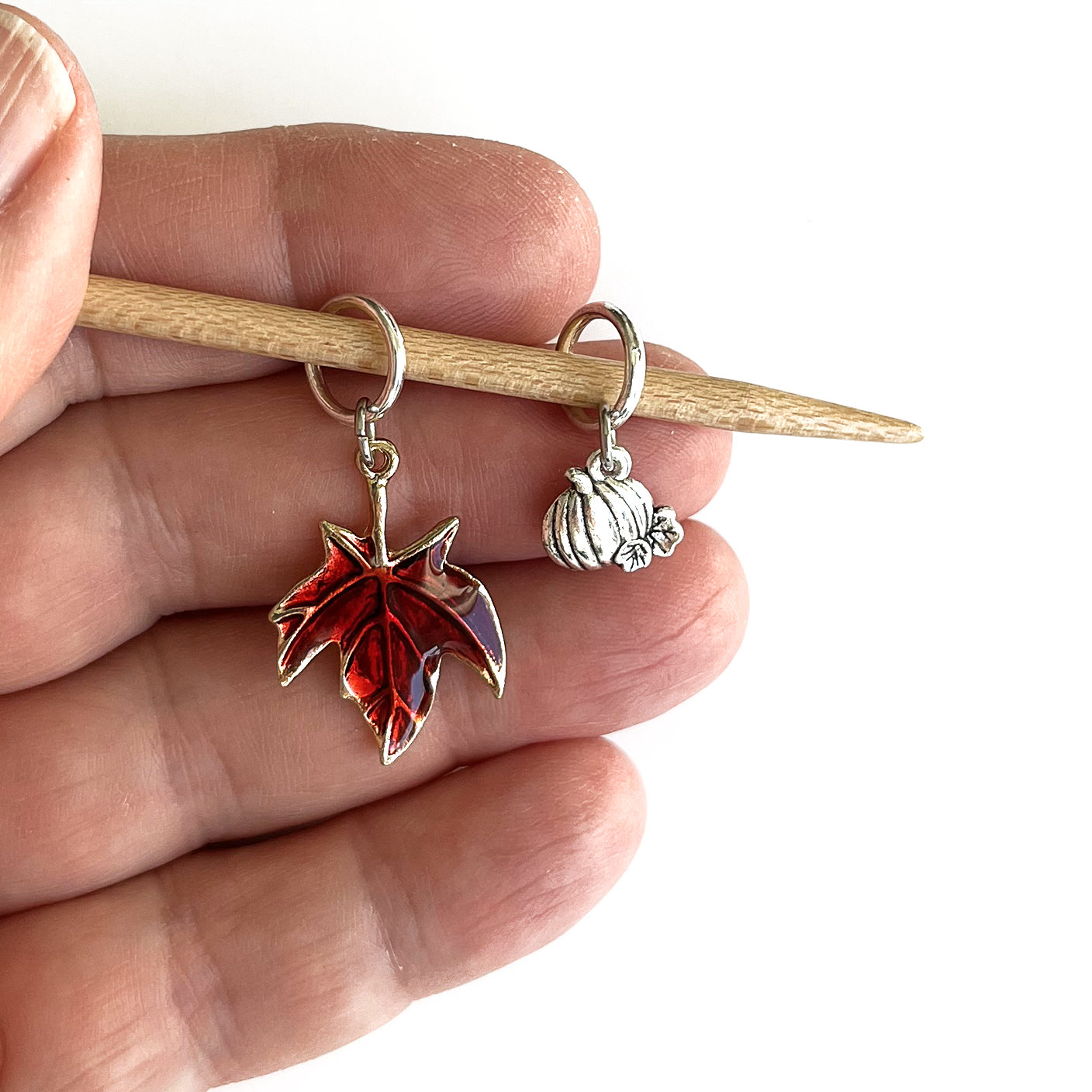 One red enamel maple leaf knitting stitch marker and one silver alloy pumpkin knitting stitch marker on a bamboo knitting needle held in a hand, made and sold by Pretty Warm Designs