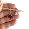 One red enamel maple leaf knitting stitch marker and one silver alloy pumpkin knitting stitch marker on a bamboo knitting needle held in a hand, made and sold by Pretty Warm Designs