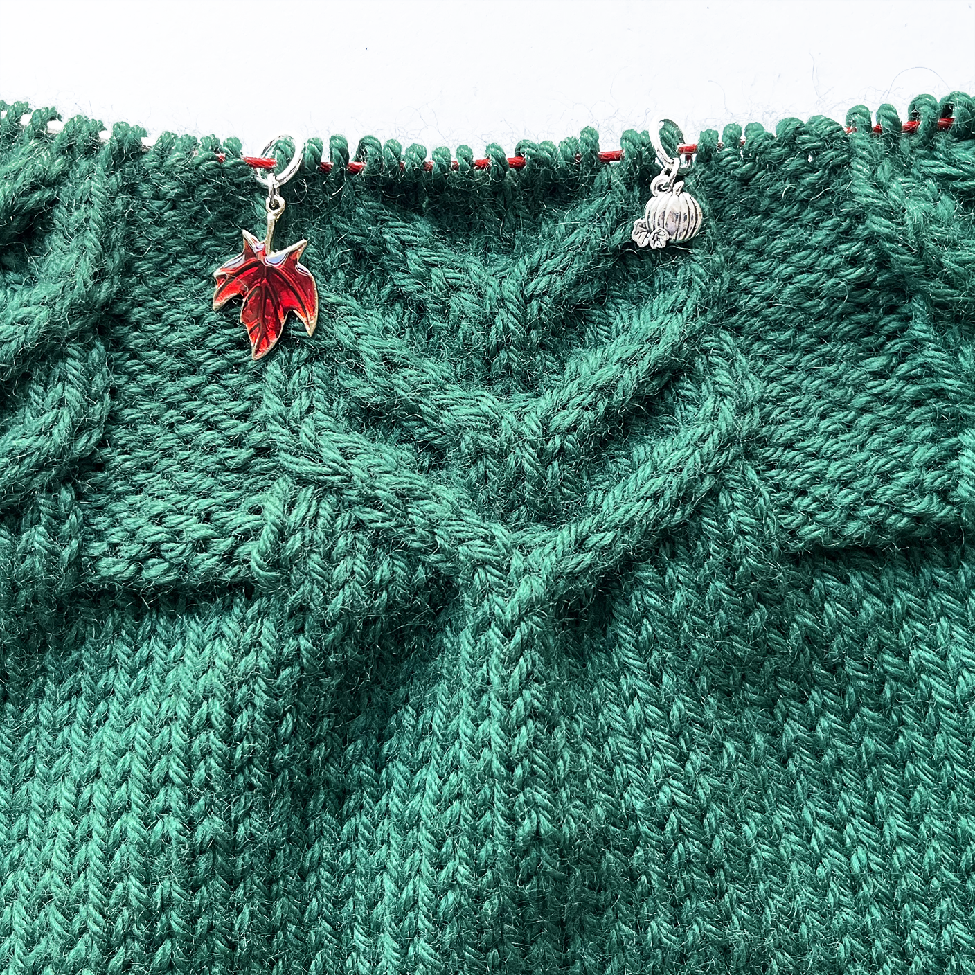 One red and gold Maple leaf knitting stitch marker and one silver alloy pumpkin knitting stitch marker threaded on a red knitting needle cable laying on a green knitting background sold by Pretty Warm Designs Inc.