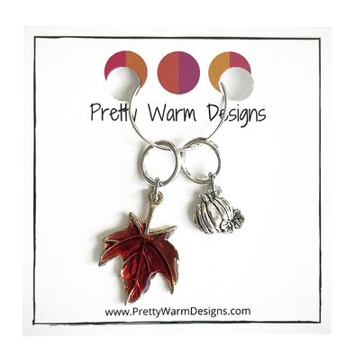 Two Fall-themed knitting ring stitch markers, one red enamel maple leaf and one silver toned pumpkin attached with a silver hoop to cardstock with Pretty Warm Designs text and logo