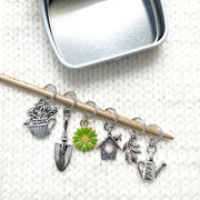 Set of one green and yellow enamel daisy and five antiqued silver alloy gardening themed snag free ring stitch markers for knitting placed on bamboo knitting needle and decorative storage tin on white knitted background by Pretty Warm Designs Inc