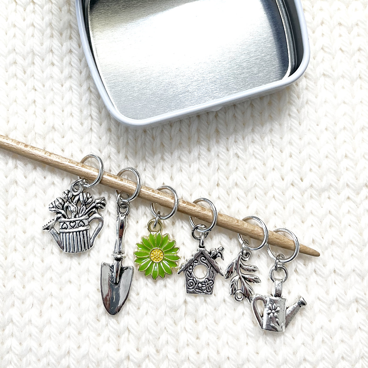 Set of one green and yellow enamel daisy and five antiqued silver alloy gardening themed snag free ring stitch markers for knitting placed on bamboo knitting needle and decorative storage tin on white knitted background by Pretty Warm Designs Inc