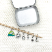 Set of one two-toned green and silver enamel tiny camper and five antiqued silver camping themed stitch markers on bamboo knitting needle with storage tin on white knitted background for knitting by Pretty Warm Designs
