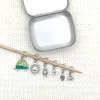 Set of one two-toned green and silver enamel tiny camper and five antiqued silver camping themed stitch markers on bamboo knitting needle with storage tin on white knitted background for knitting by Pretty Warm Designs