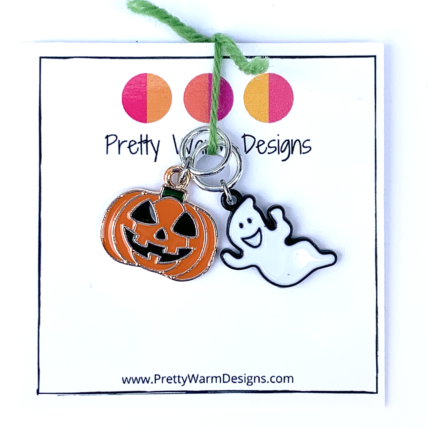 Two Hallowe'en-themed knitting ring stitch markers, one orange, black and green enamel jack-o-lantern and one white and black enamel ghost attached with green yarn to cardstock with Pretty Warm Designs text and logo