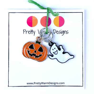 Two Hallowe'en-themed knitting ring stitch markers, one orange, black and green enamel jack-o-lantern and one white and black enamel ghost attached with green yarn to cardstock with Pretty Warm Designs text and logo