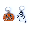Two Hallowe'en-themed knitting ring stitch markers, one orange, black and green enamel jack-o-lantern and one white and black enamel ghost by Pretty Warm Designs