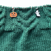 One orange, black and green enamel jack-o-lantern knitting stitch marker and one white and black enamel ghost knitting stitch marker threaded on a red knitting needle cable laying on a green knitted background sold by Pretty Warm Designs Inc.