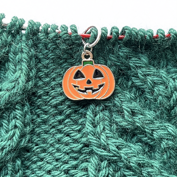One orange, black and green enamel jack-o-lantern knitting stitch marker threaded on a red knitting needle cable laying on a green knitted background sold by Pretty Warm Designs Inc.