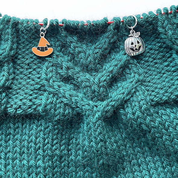 One Halloween themed orange and black enamel witch hat knitting stitch marker and one silver toned jack-o-lantern stitch marker threaded on a red knitting needle cable on top of a green knitted background and sold by Pretty Warm Designs Inc.