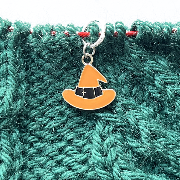 One Halloween themed orange and black enamel witch hat knitting stitch marker threaded on a red knitting needle cable on top of a green knitted background and sold by Pretty Warm Designs Inc.
