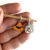 Two Hallowe'en-themed knitting ring stitch markers, one orange and black enamel witch hat and one silver toned jack-o-lantern on bamboo needle held by a hand, made and sold by Pretty Warm Designs