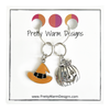 Two Hallowe'en-themed knitting ring stitch markers, one orange and black enamel witch hat and one silver toned jack-o-lantern attached with silver hoop ring to cardstock with Pretty Warm Designs text and logo