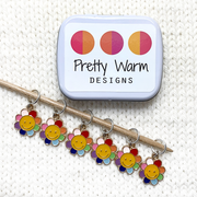 Set of six yellow and rainbow petal enamel happy face flower charms snag free ring stitch markers on bamboo knitting needle resting on white knitted fabric with open tin for knitting by Pretty Warm Designs