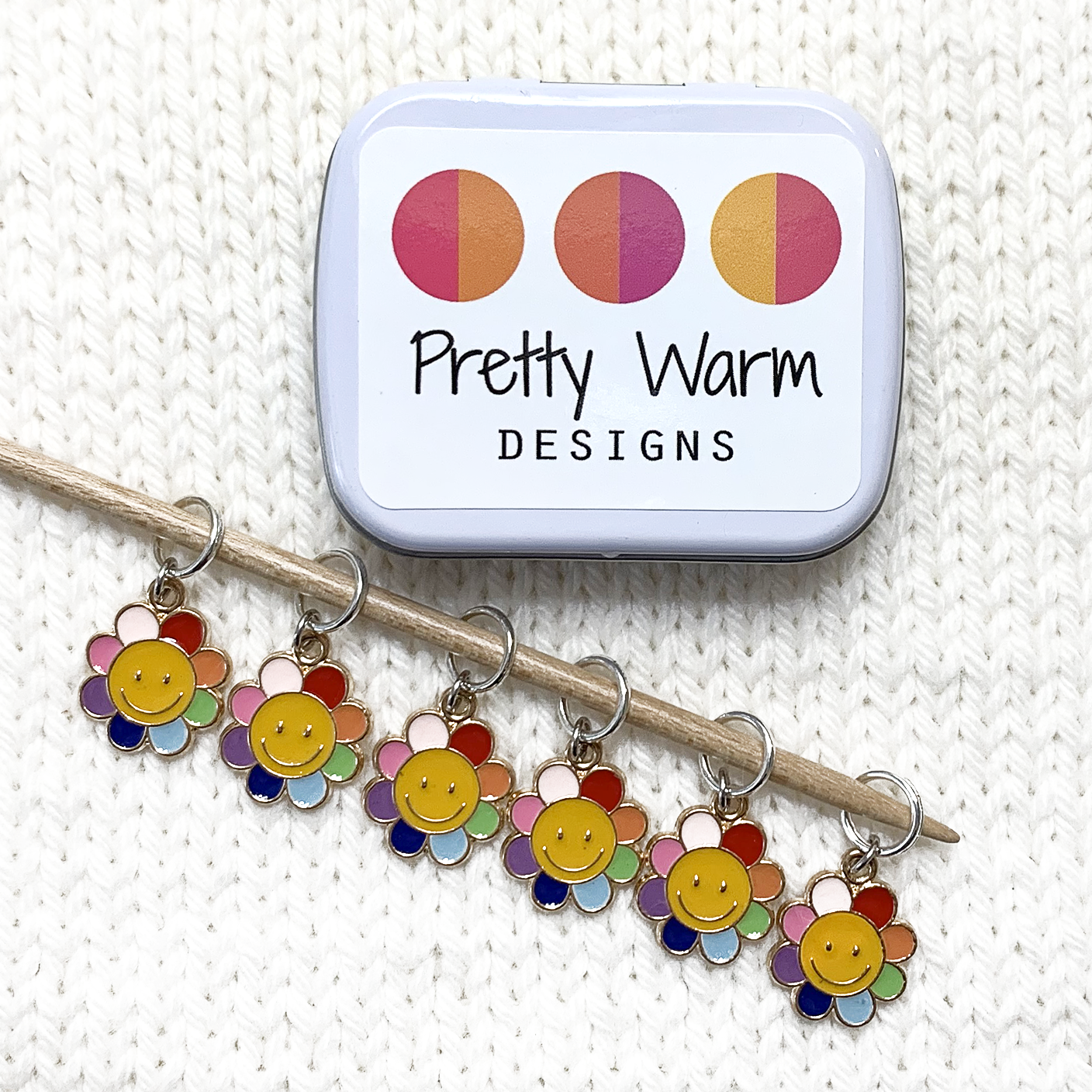 Set of six yellow and rainbow petal enamel happy face flower charms snag free ring stitch markers on bamboo knitting needle resting on white knitted fabric with open tin for knitting by Pretty Warm Designs