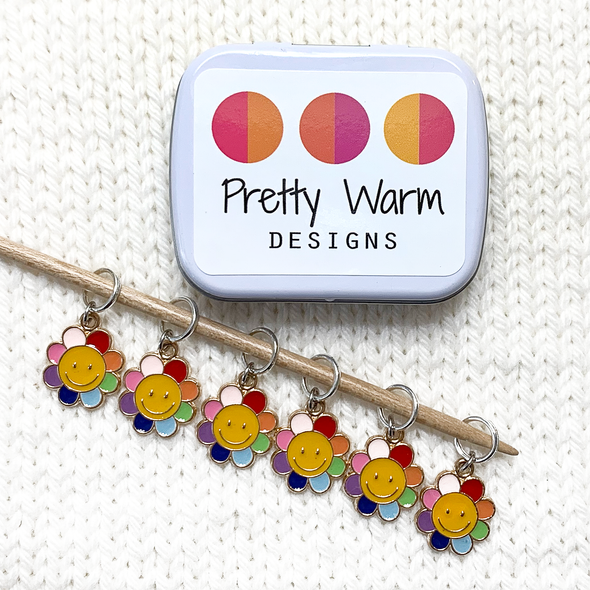 Set of six yellow and rainbow petal enamel happy face flower charms snag free ring stitch markers on bamboo knitting needle resting on white knitted fabric with open tin for knitting by Pretty Warm Designs