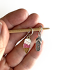 Two summer-themed knitting ring stitch markers, one enamel pink and brown ice cream cone and one silver toned flip flop on a bamboo knitting needle held by a hand, made and sold by Pretty Warm Designs