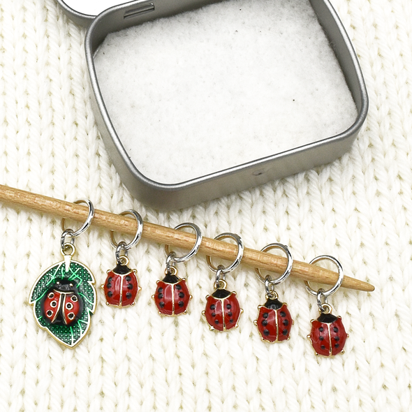 Set of five red and black enamel ladybug and one red, black and green ladybug on a leaf charms snag free ring stitch markers on knitting needle with open tin for knitting by Pretty Warm Designs