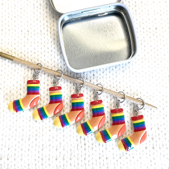 Set of six large rainbow coloured resin charm stitch markers on knitting needle and decorative tin sitting on white knitted background by Pretty Warm Designs Inc
